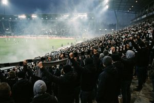 Fankurve im Stadion bei Flutlicht, Fans halten Schals hoch, leichter Rauch